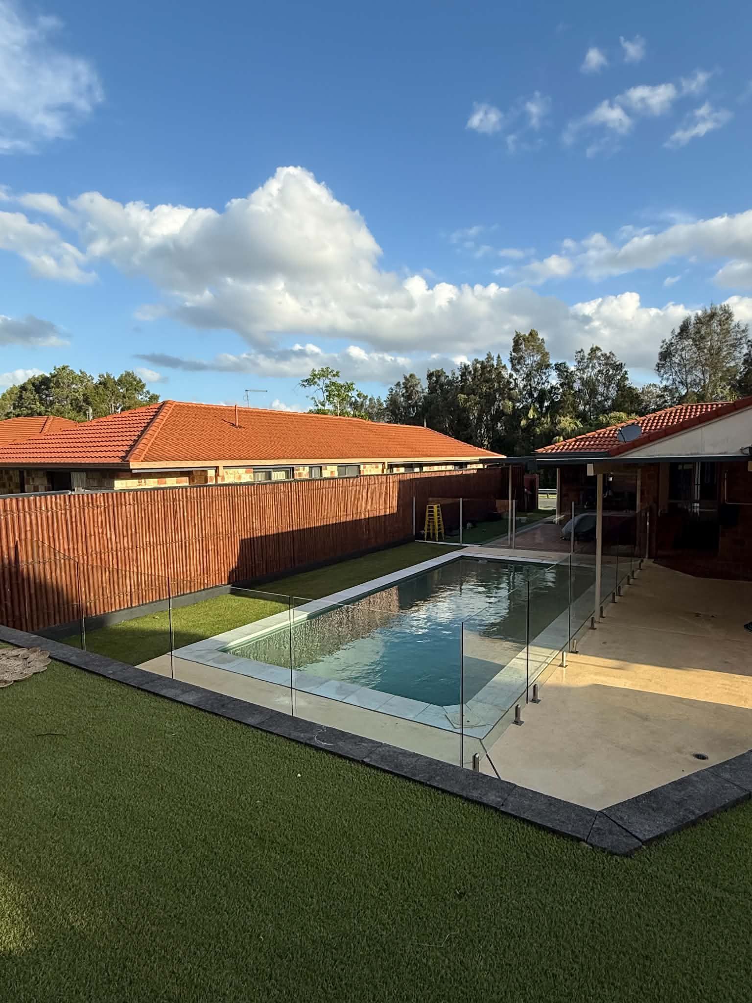 Backyard pool with glass fencing, patio, red-roofed house, and blue sky with clouds
