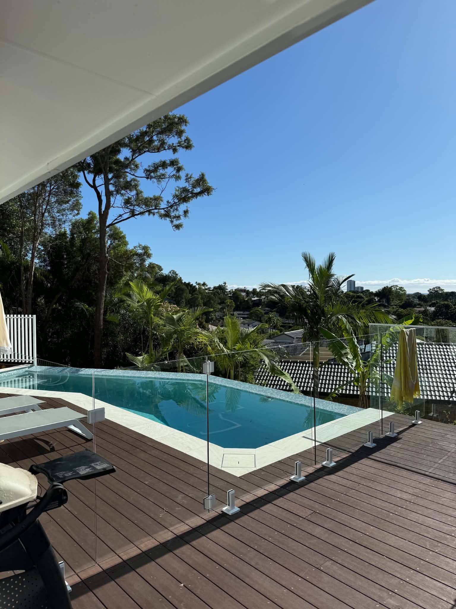 Infinity pool beside a wooden deck, with lush greenery and blue sky beyond the glass railing.