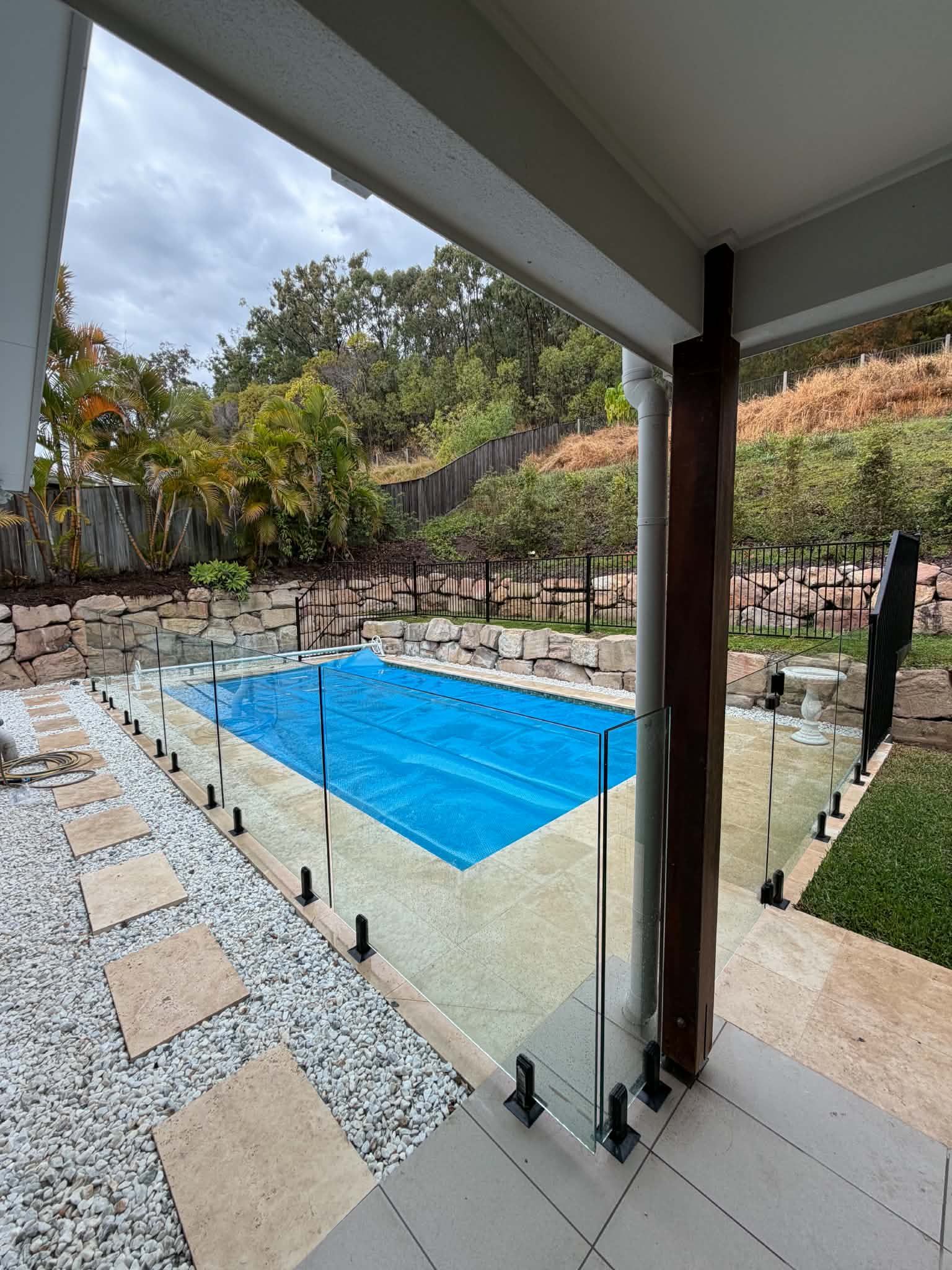 Covered patio overlooking a blue-tiled pool with glass railing and garden fencing