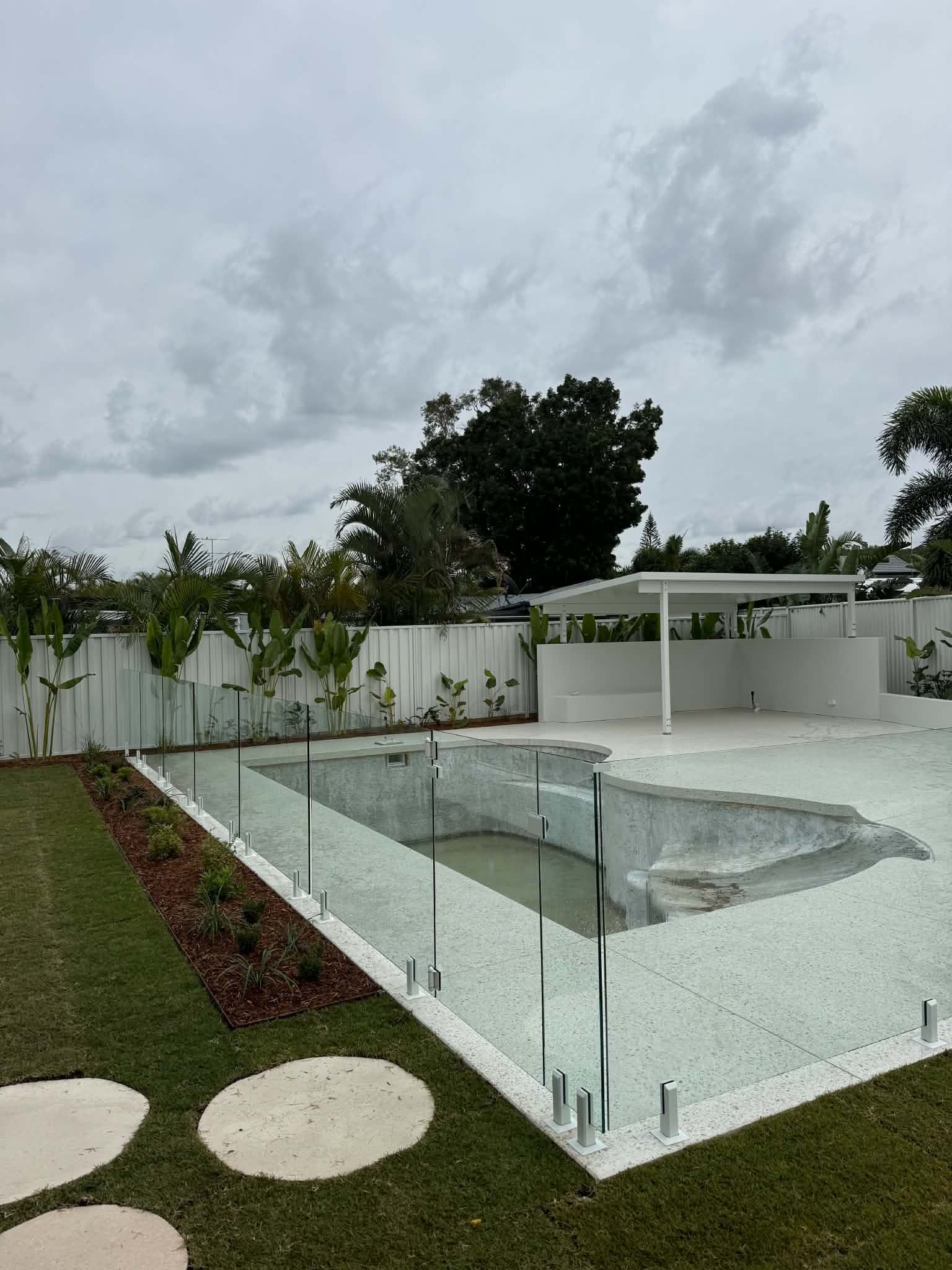 Modern backyard with a glass-fenced in-ground pool, white patio, and cloudy sky