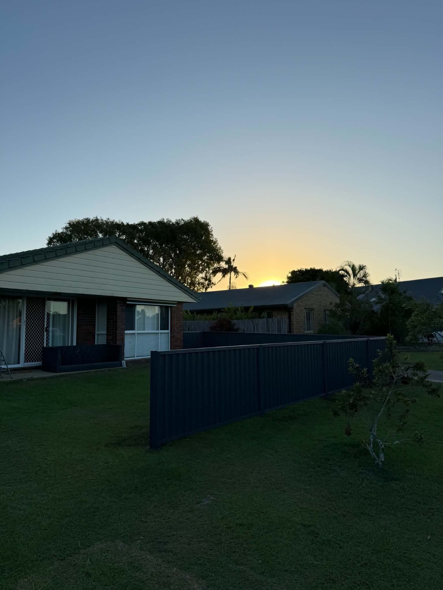 Dusk over a suburban house and lawn, with a dark fence silhouetted against the sunset.