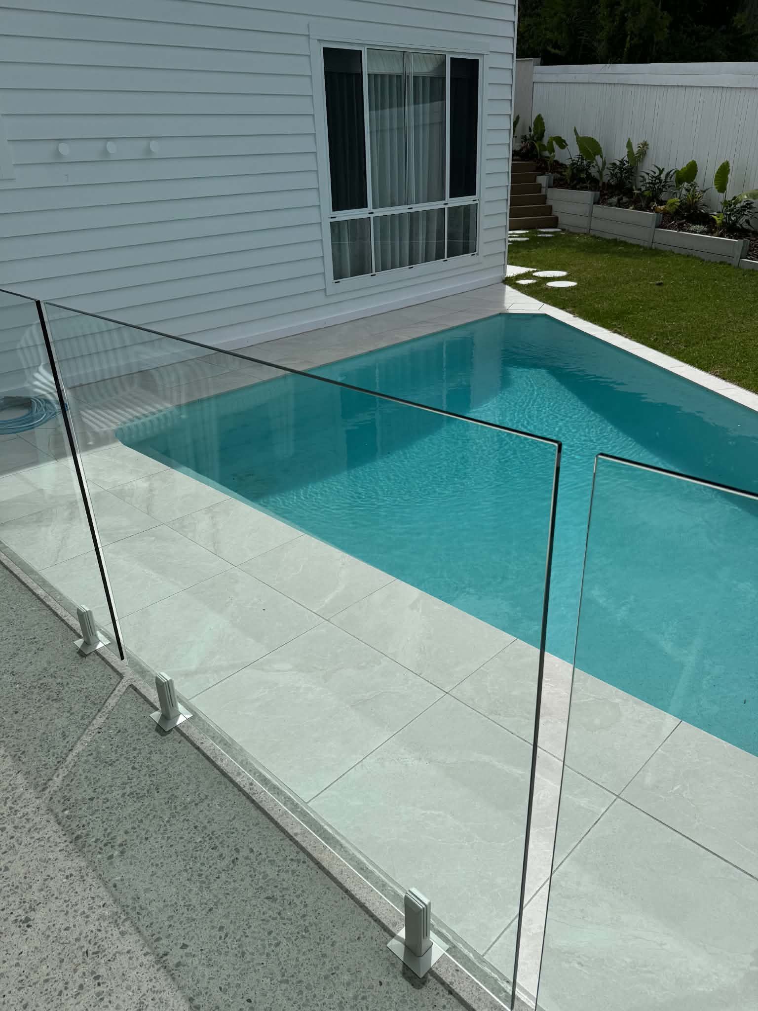 Glass fence around a turquoise backyard pool beside a white house and lawn.