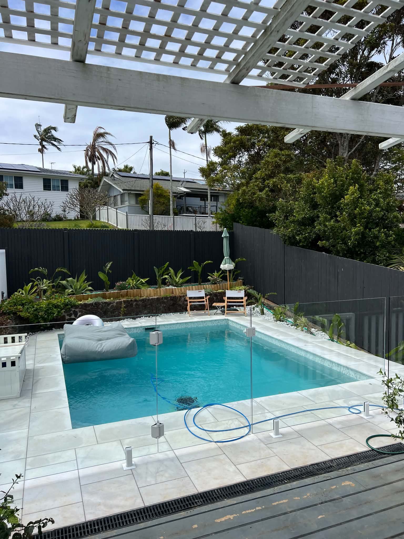 Backyard pool with turquoise water, white patio tiles, gray fence, and pergola overhead