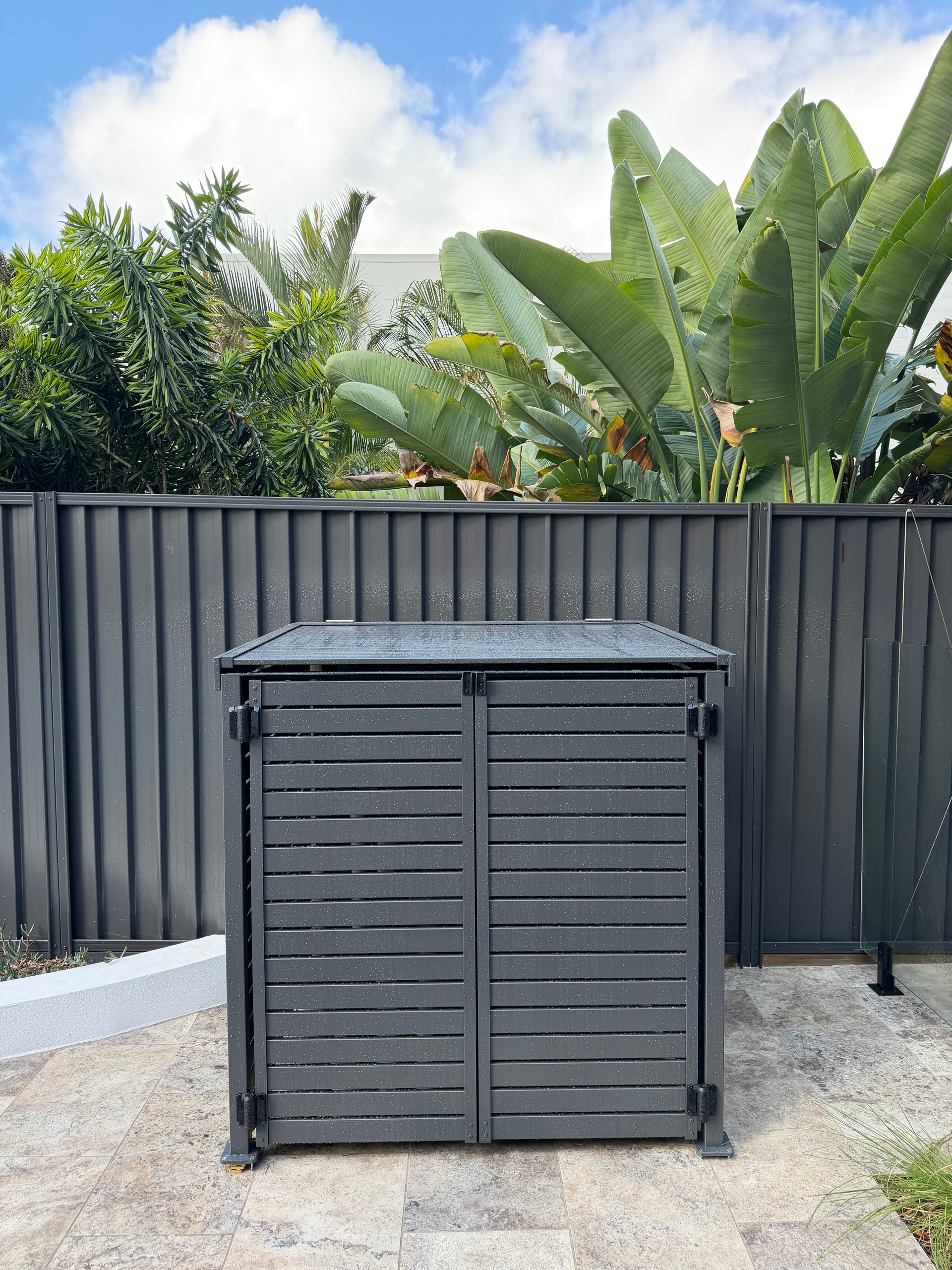 Black outdoor storage cabinet on a gravel patio in front of a gray fence and lush green plants