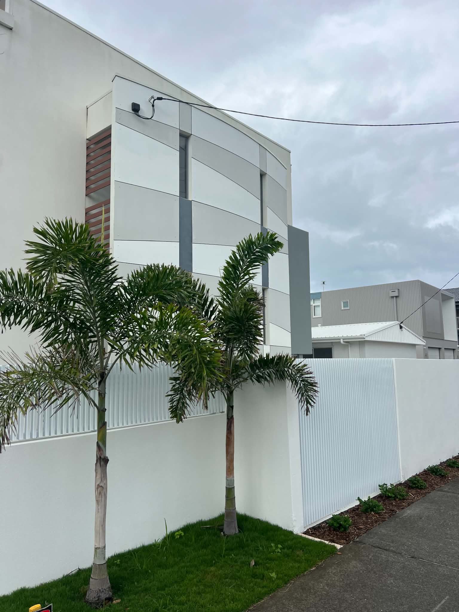 White modern building behind a white fence, with small trees and a paved roadside under cloudy sky
