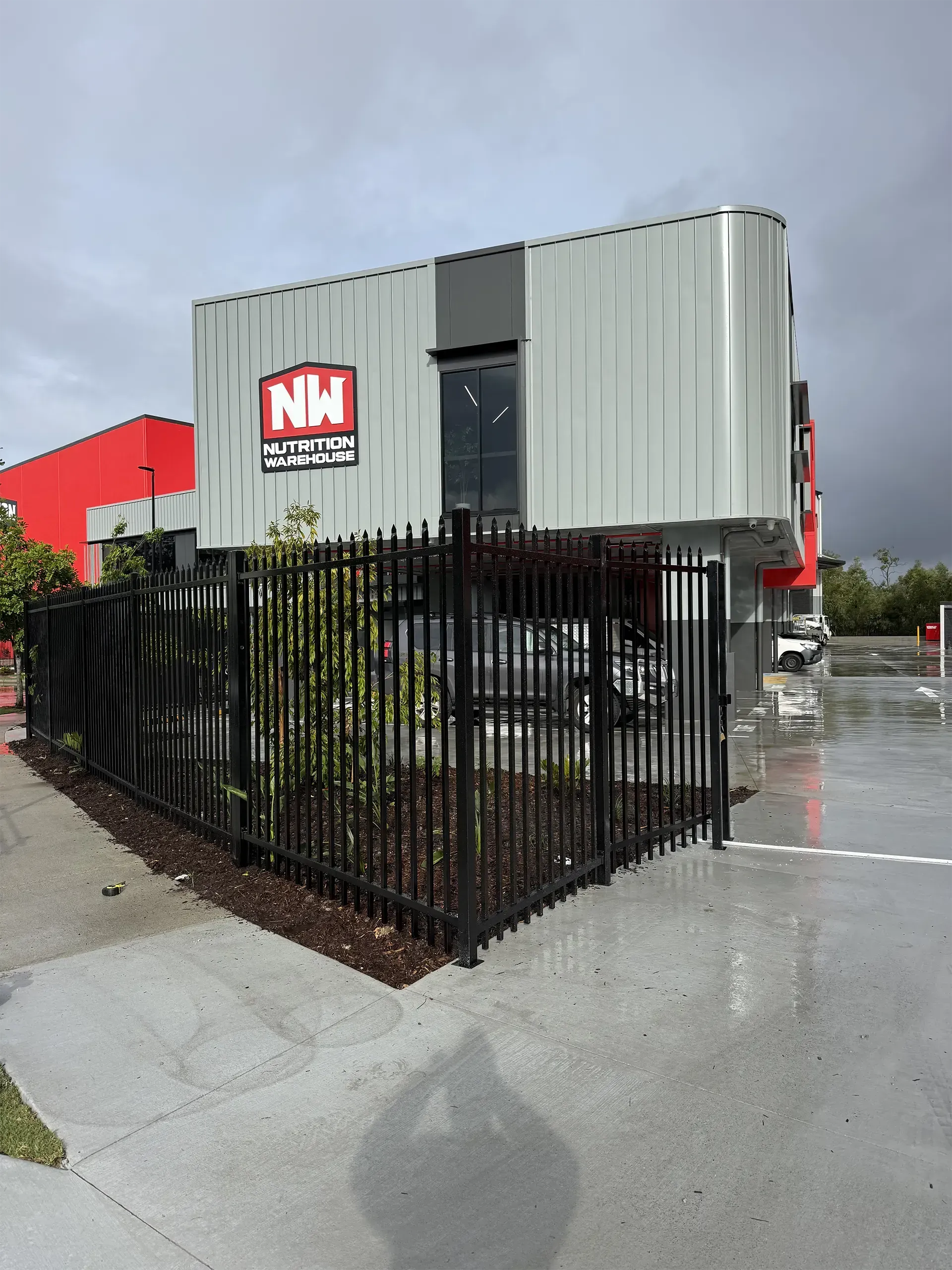 Black fenced lot with a gray modular building and red N&M sign on a wet street