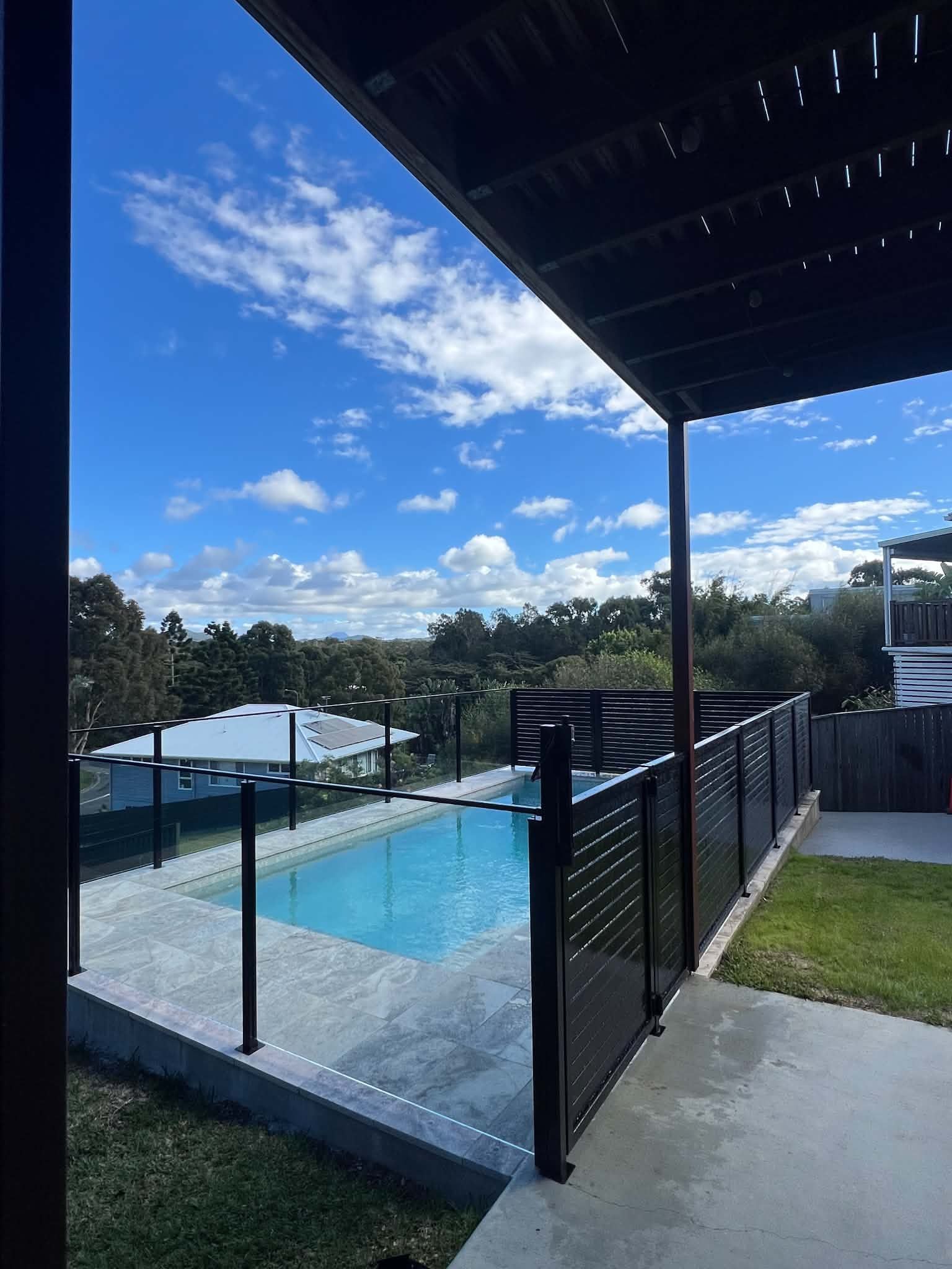 Backyard pool with black fence, blue water, and covered patio under a bright sky