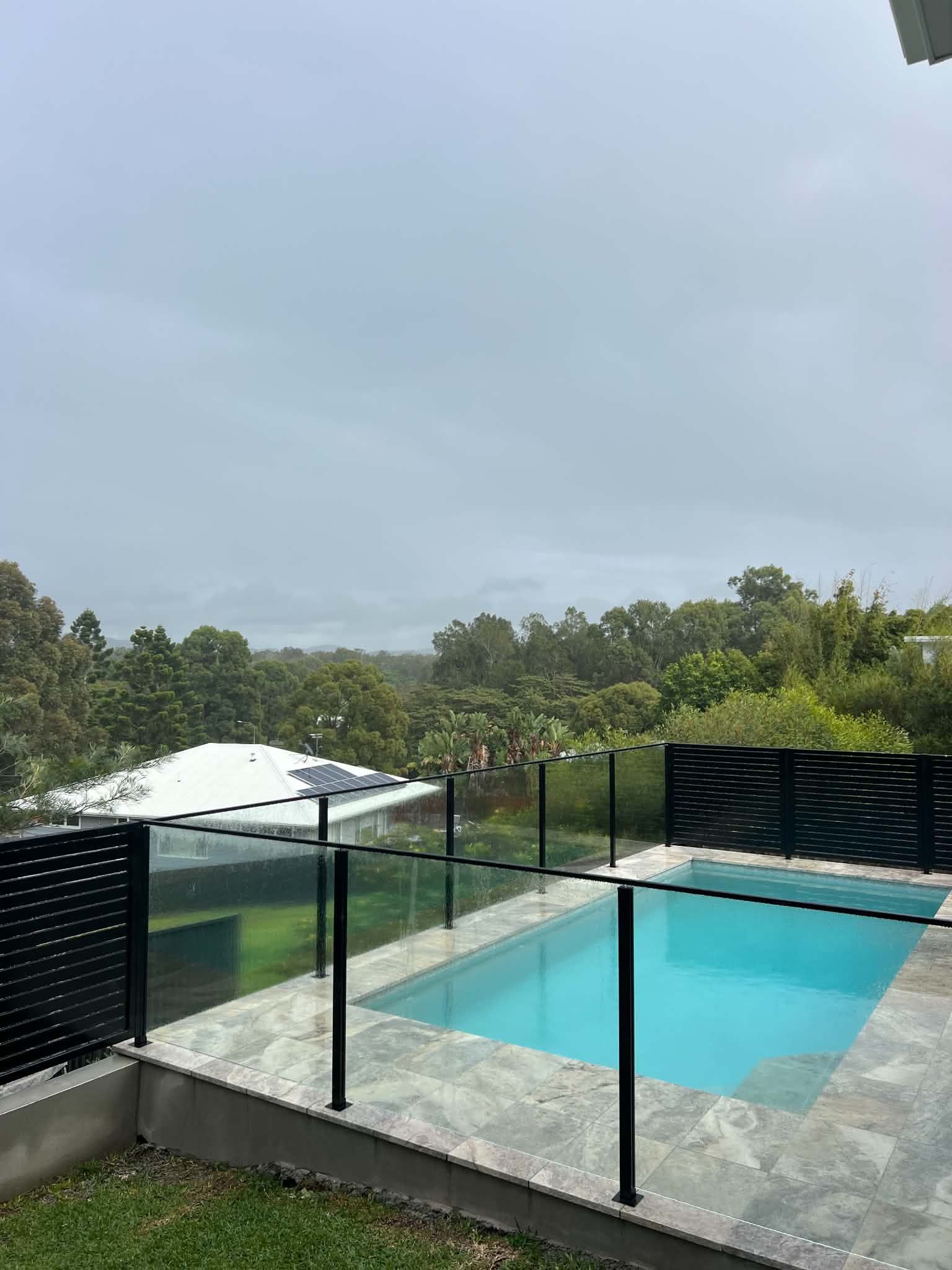 Modern backyard pool with glass railing, tiled deck, and cloudy sky overlooking trees
