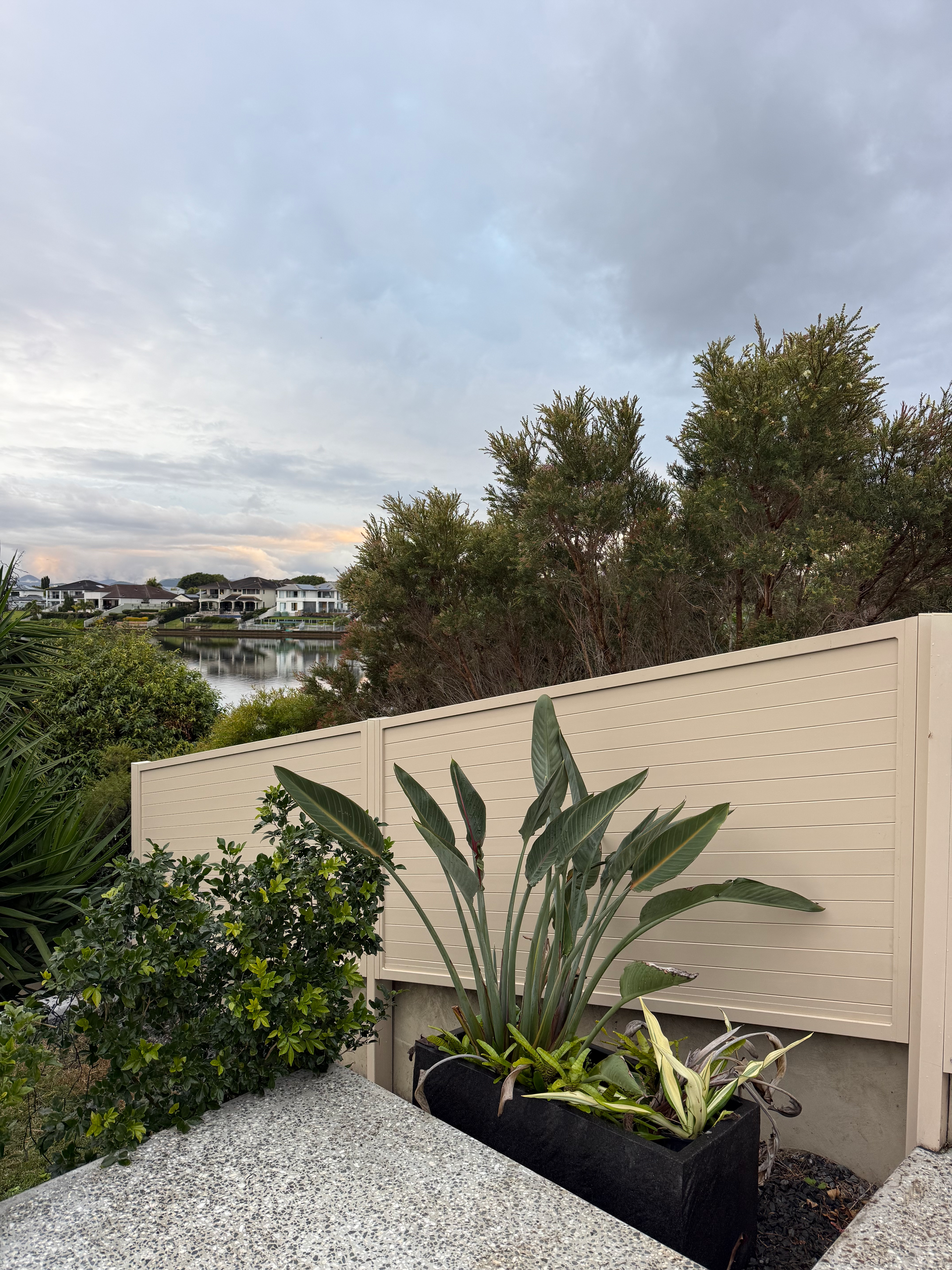 Backyard patio with potted tropical plants, gravel, and a beige fence overlooking a distant city skyline
