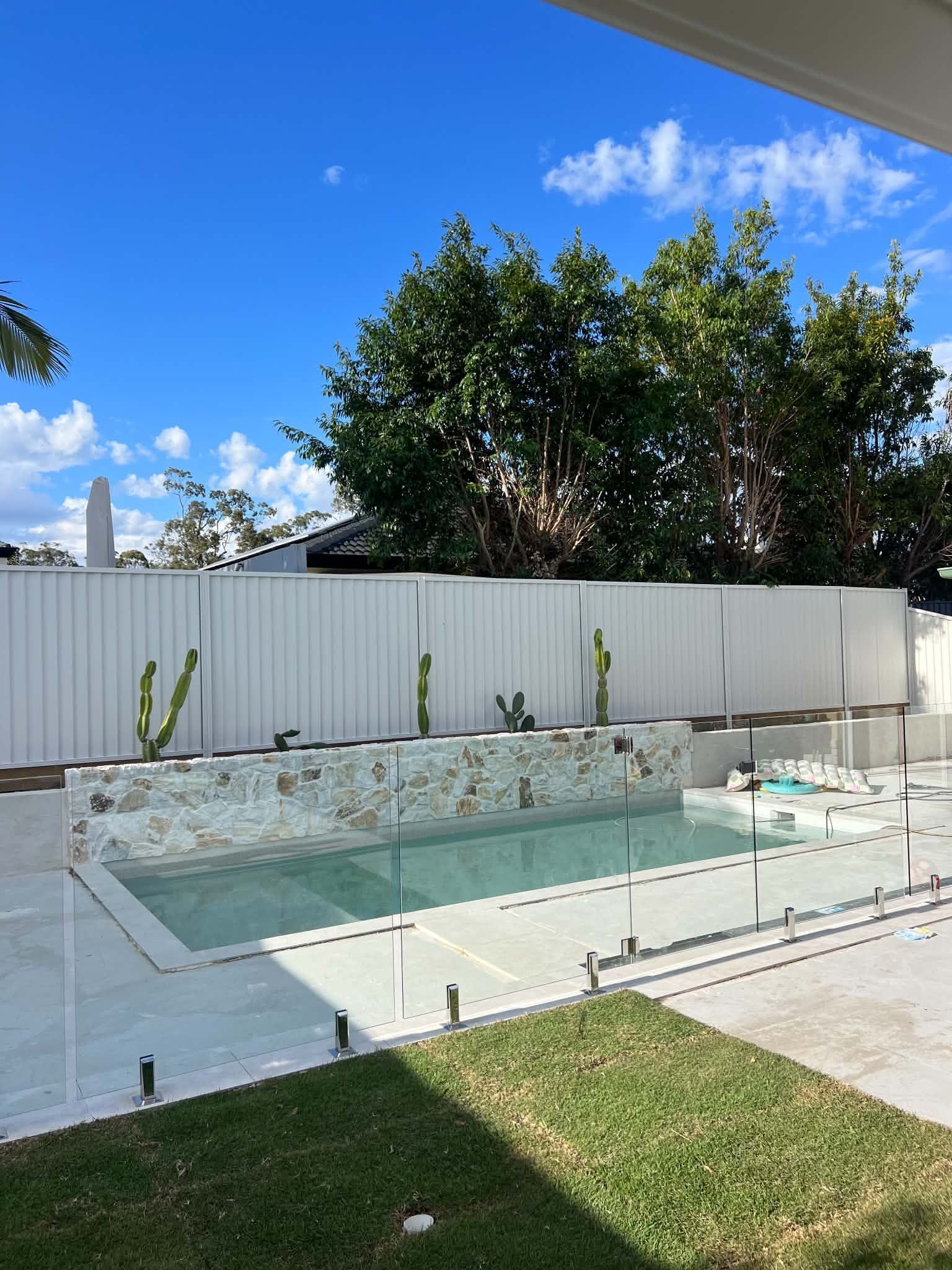 Backyard with a rectangular swimming pool, white fence, and lush green trees under a blue sky.