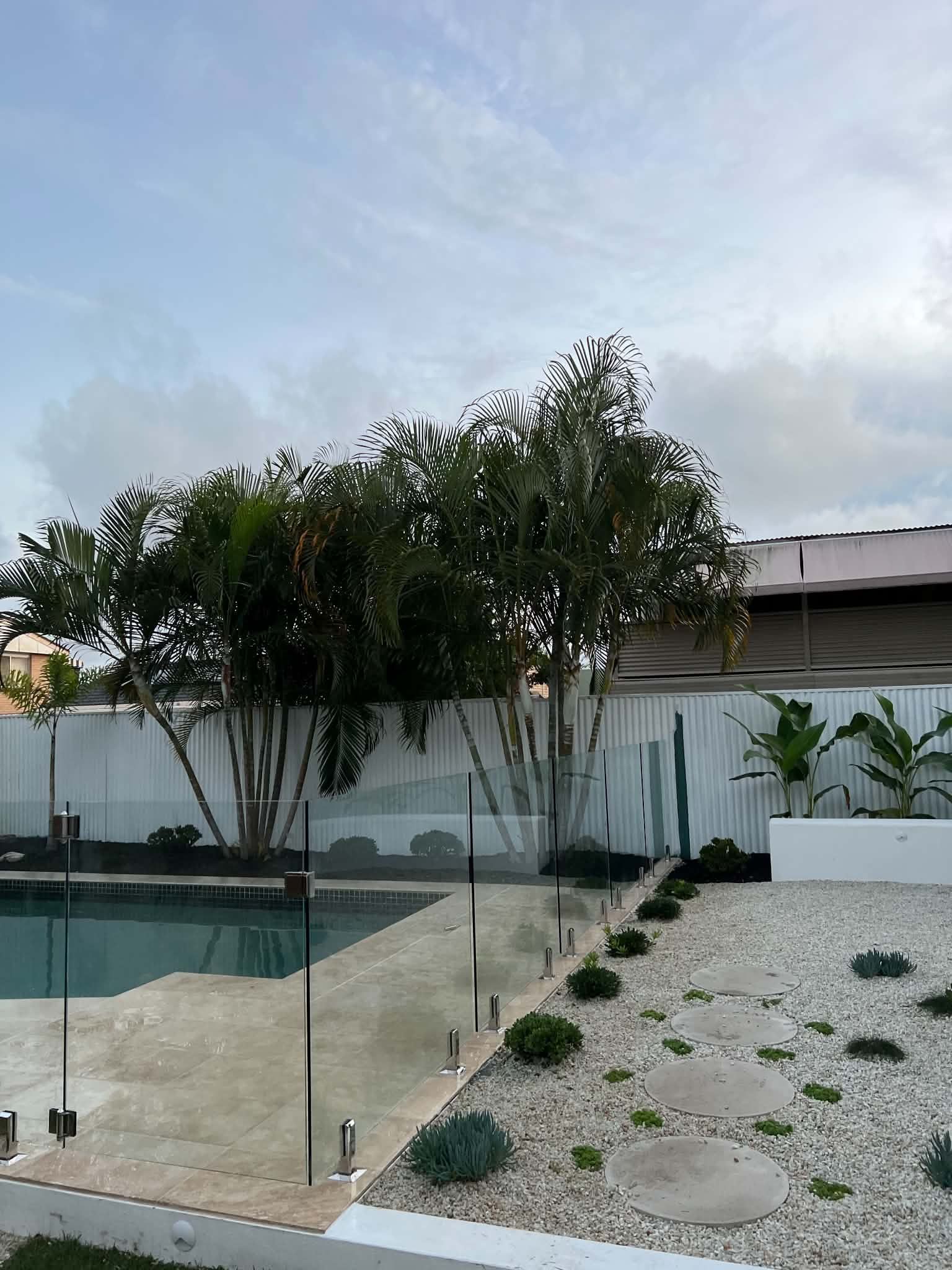 Backyard pool with glass fence, gravel landscaping, and tall trees under a cloudy sky