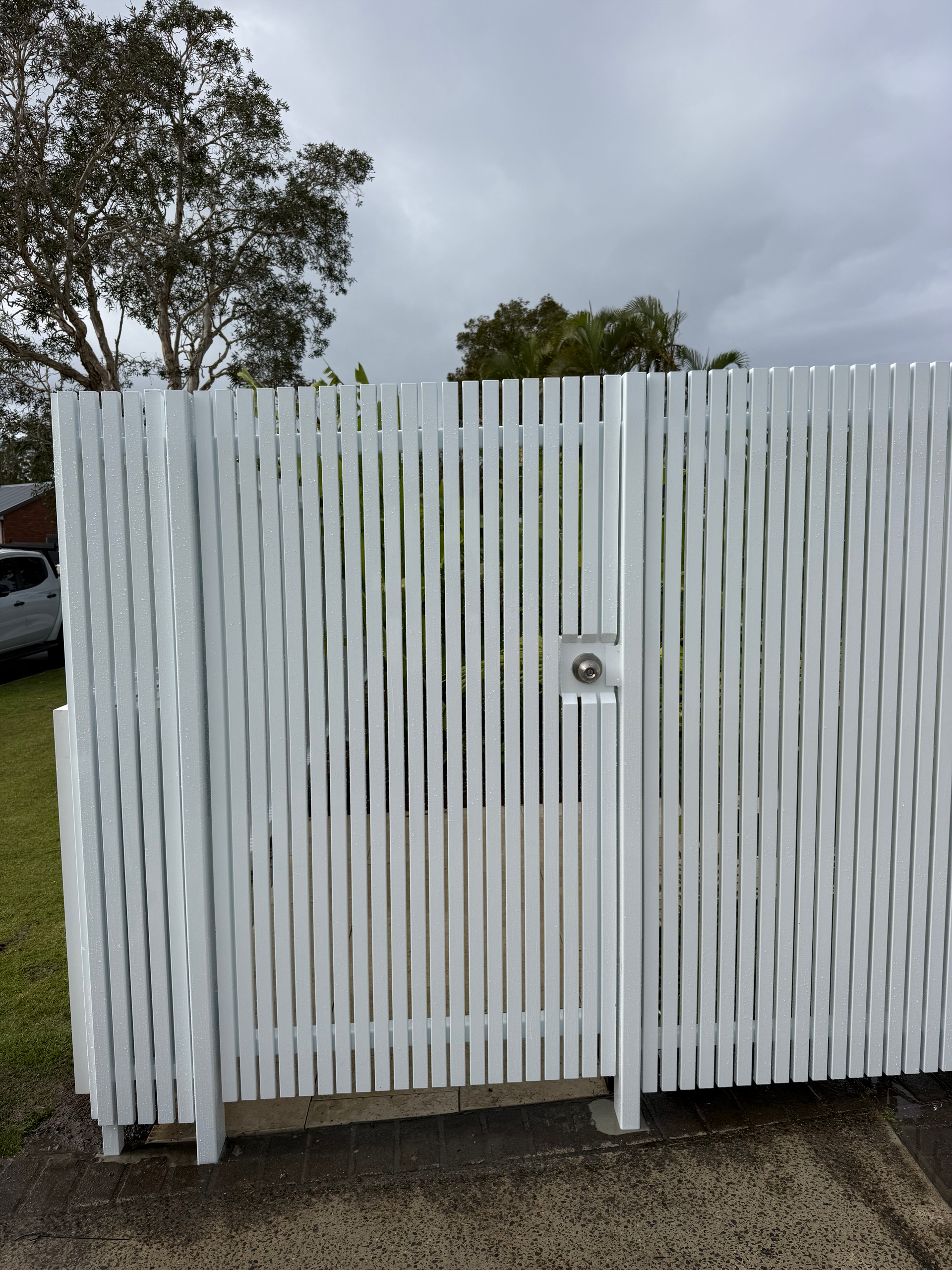 White slatted gate with a small latch, set in a driveway under a cloudy sky.