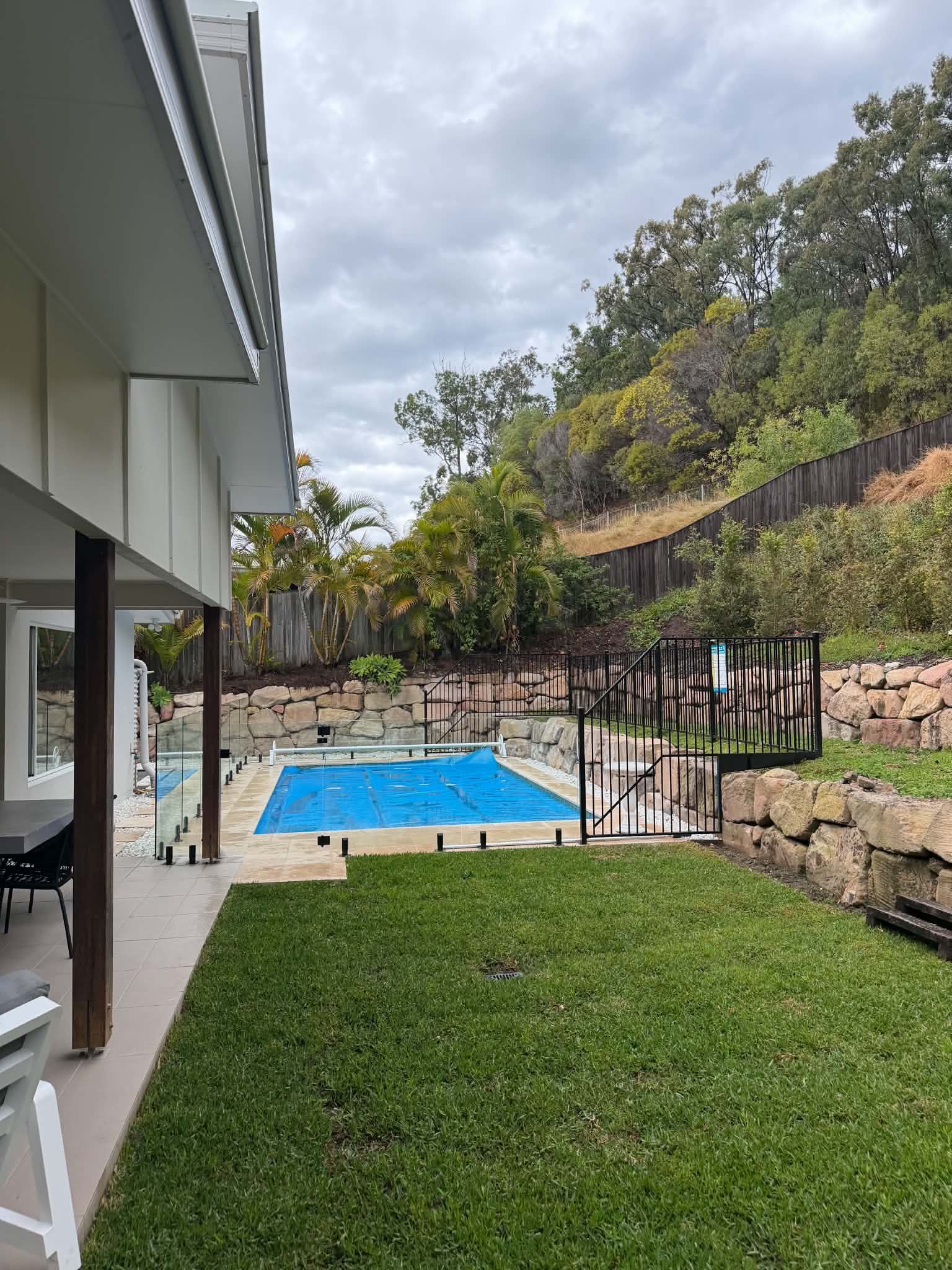 Backyard patio with green lawn, blue pool, stone retaining wall, and hillside trees under cloudy sky