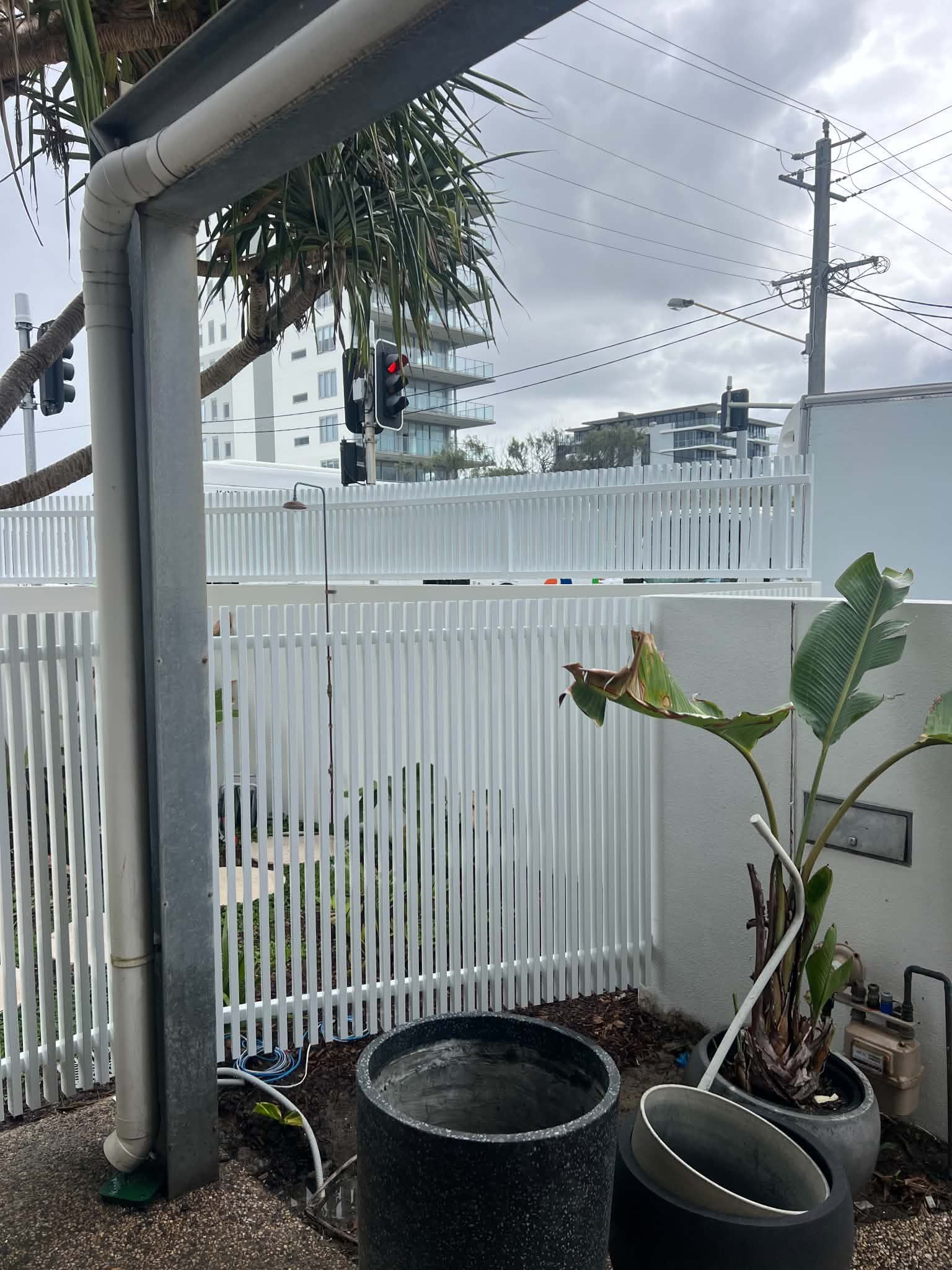 Patio corner with potted plants, a metal post, and a white fence under a cloudy sky.