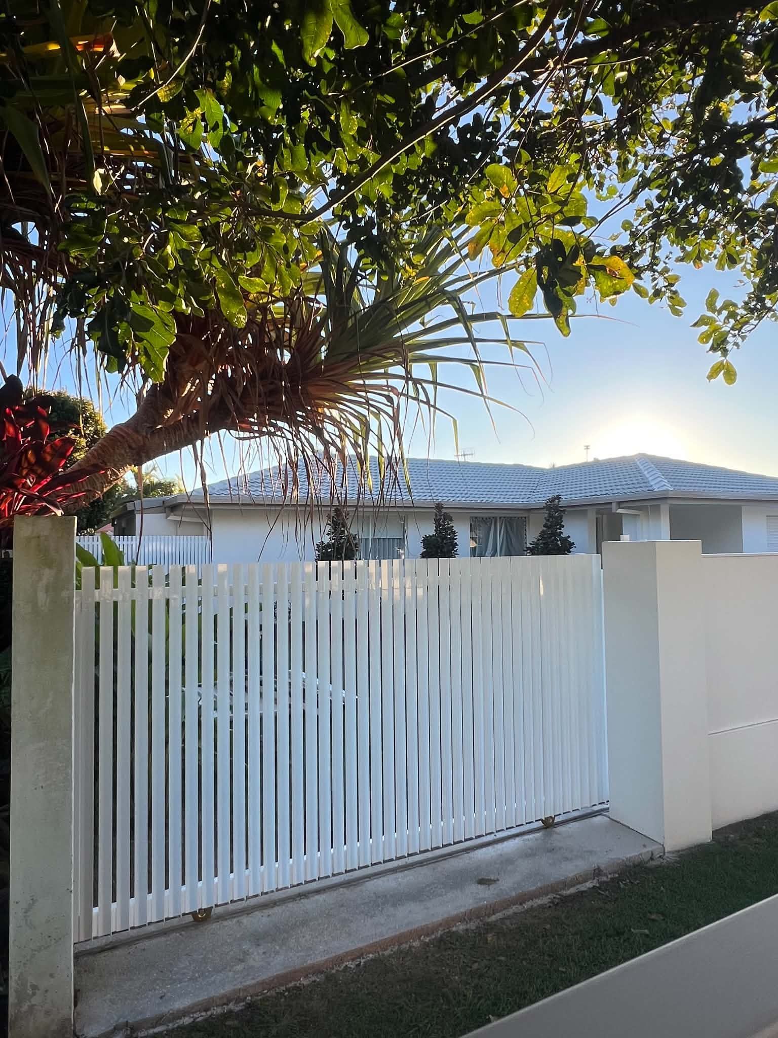 White slatted gate beside a driveway, shaded by a leafy tree, with a house and blue sky behind it