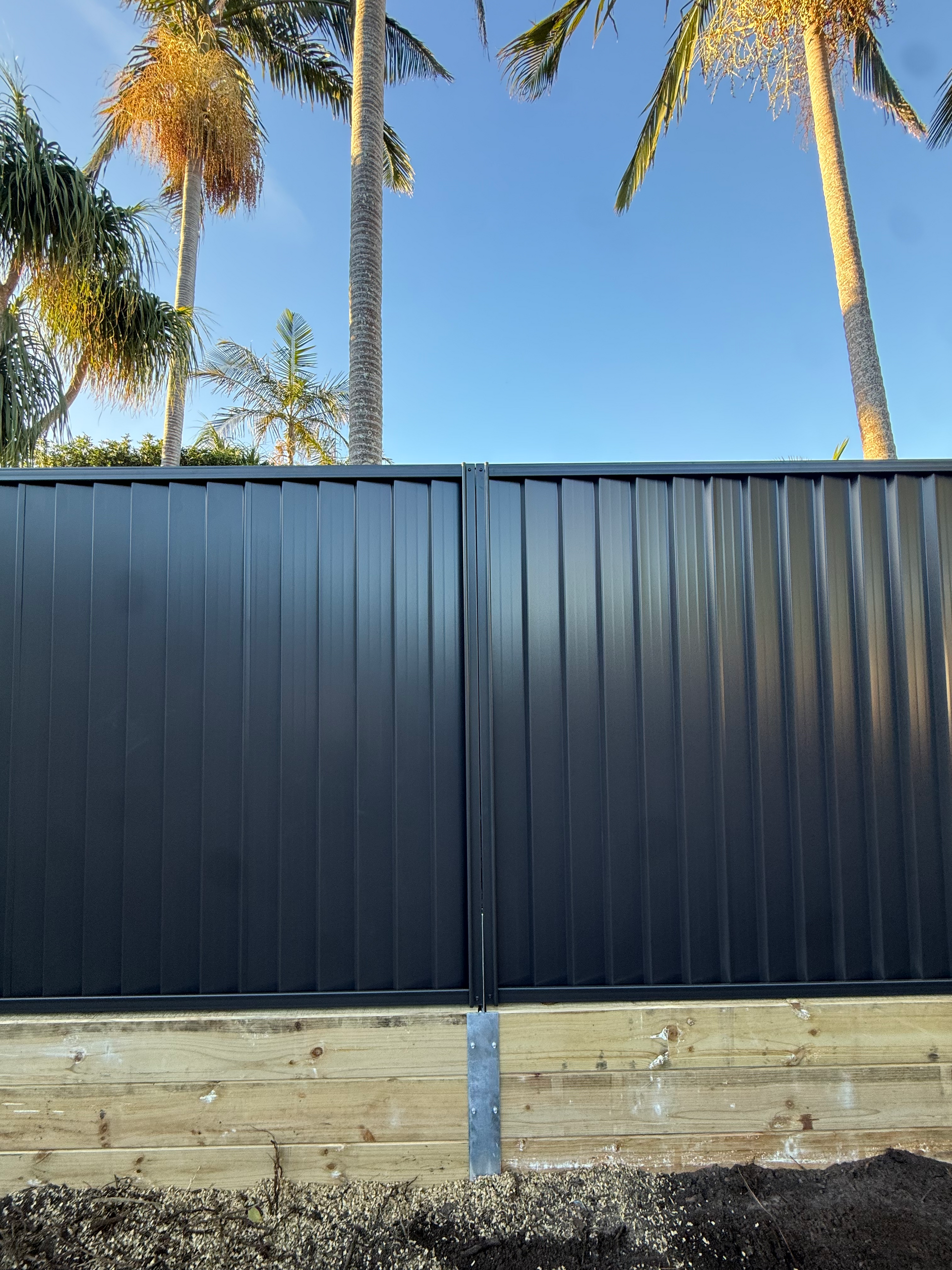 Black corrugated fence with stone base, palm trees visible above against a clear blue sky.