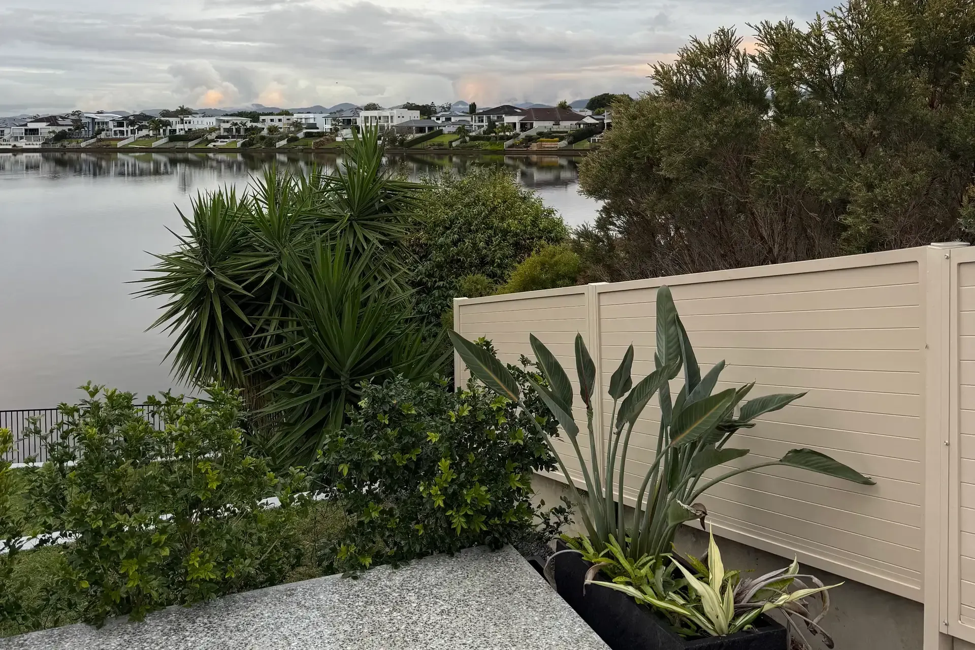 Backyard patio with potted tropical plants overlooking a calm waterfront and cloudy sky