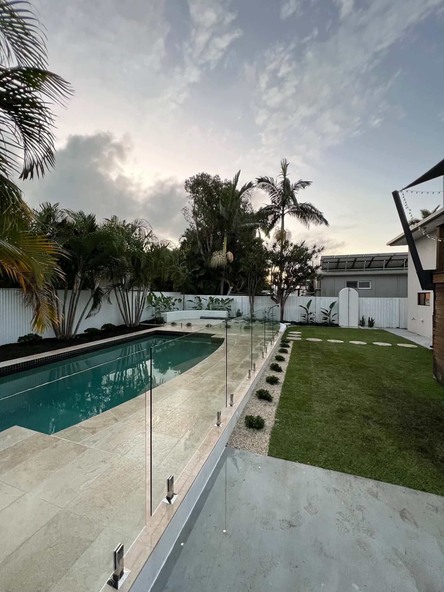 Modern backyard patio with a swimming pool, glass fence, green lawn, and palm trees at sunset