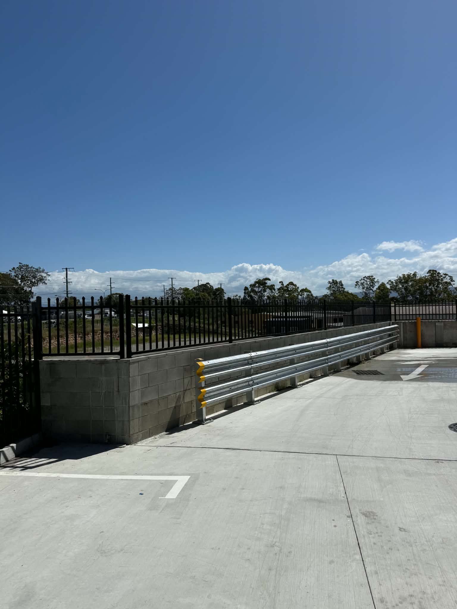 Sunny parking lot with a black metal fence, concrete wall, and open blue sky.