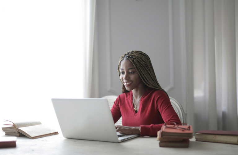 Woman in a red sweater working on a laptop at a desk by a window