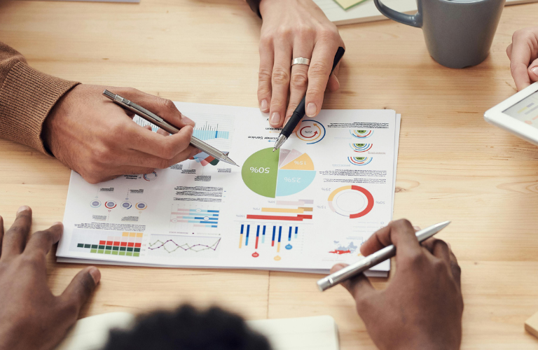Hands collaborating over a business chart with graphs, charts, and pens on a wooden table