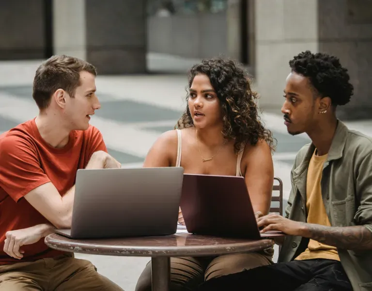 Three people talking around laptops at a small outdoor table in a courtyard