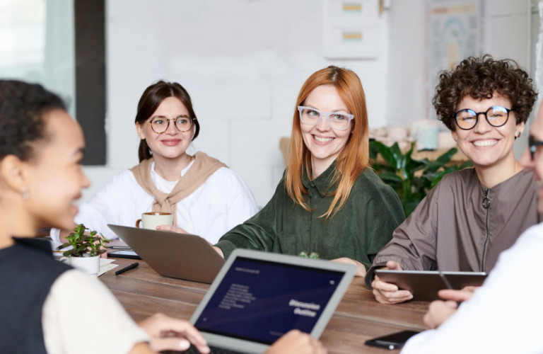 Team meeting with four people around a table, smiling and using laptops in a bright office.