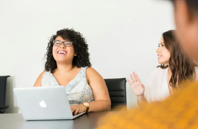 Two people talking in a meeting room, one laughing behind a laptop, another gesturing beside them.