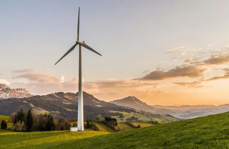 Wind turbine on a green hillside at sunset with mountains and clouds in the background