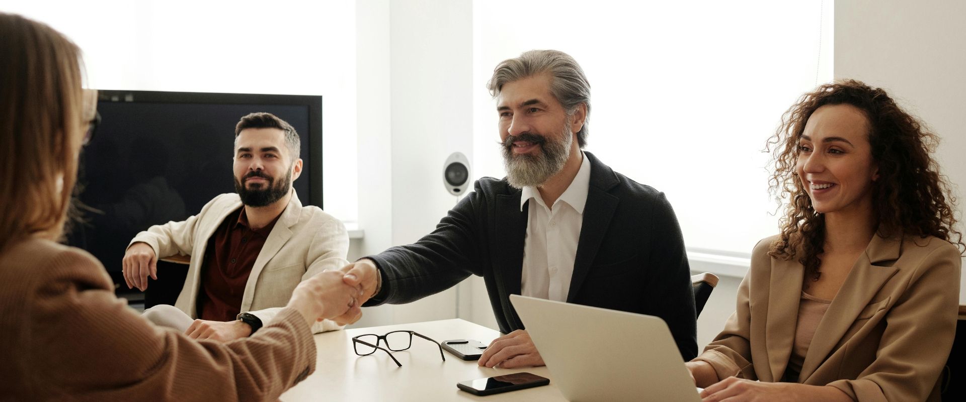 Business meeting with three people at a table, shaking hands, with a laptop and glasses in a bright office.