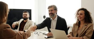 Business meeting with three people at a table, shaking hands, with a laptop and glasses in a bright office.