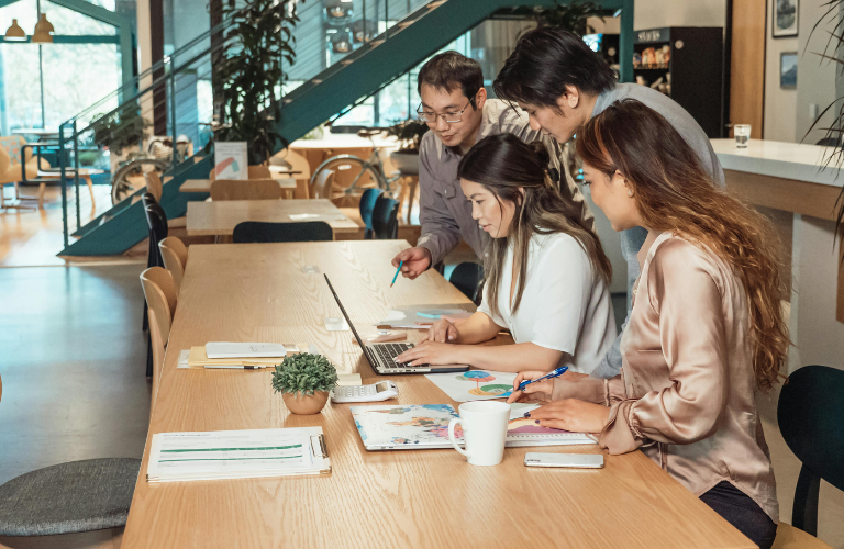 People collaborating at a long table in a bright office, writing and using laptops.