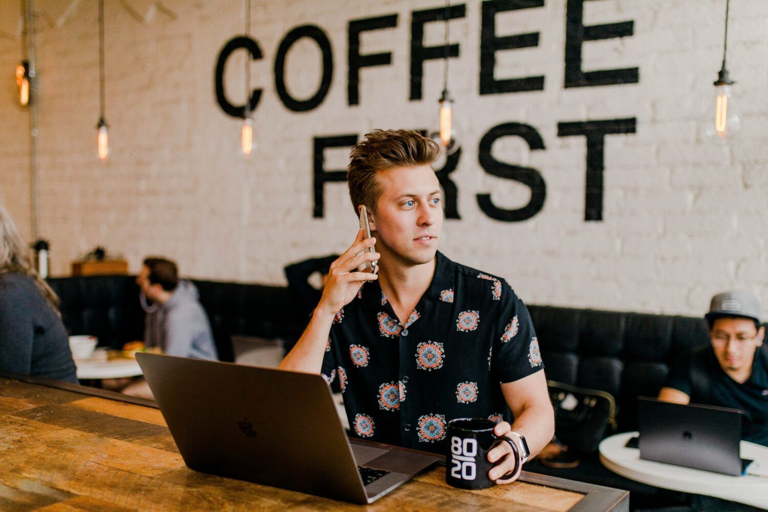 Person working on a laptop in a coffee shop with “COFFEE FIRST” on the wall