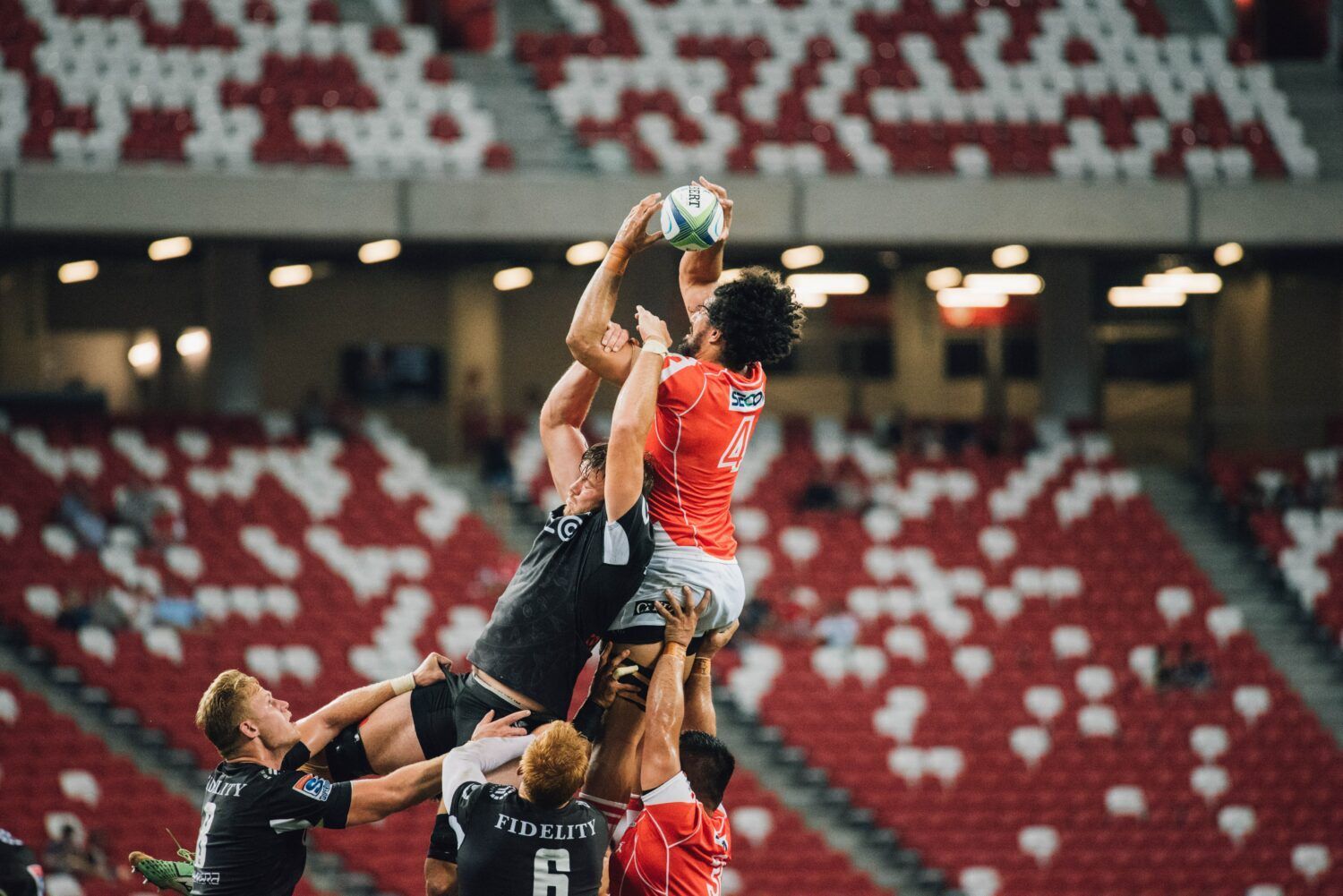 Rugby players leap for a contested ball during a match in a stadium with red-and-white seats.