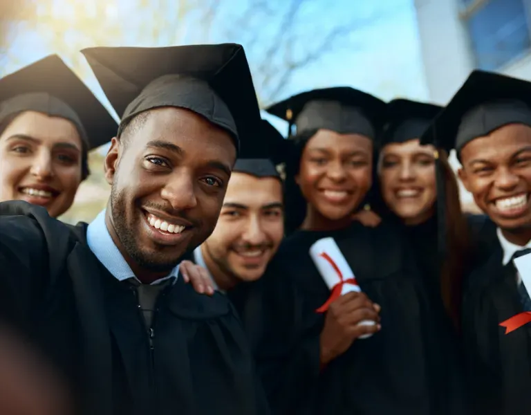 Smiling graduates in caps and gowns posing outdoors, one holding a rolled diploma.