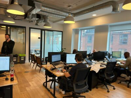 Open-plan office with people working at desks under hanging lights near large windows.