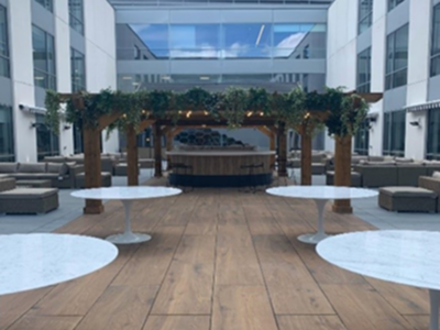 Modern indoor atrium with wood floor, round white tables, gray seating, and a decorated kiosk under glass skylights