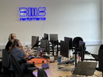 Office workspace with several monitors and two people working at desks under a blue wall logo.