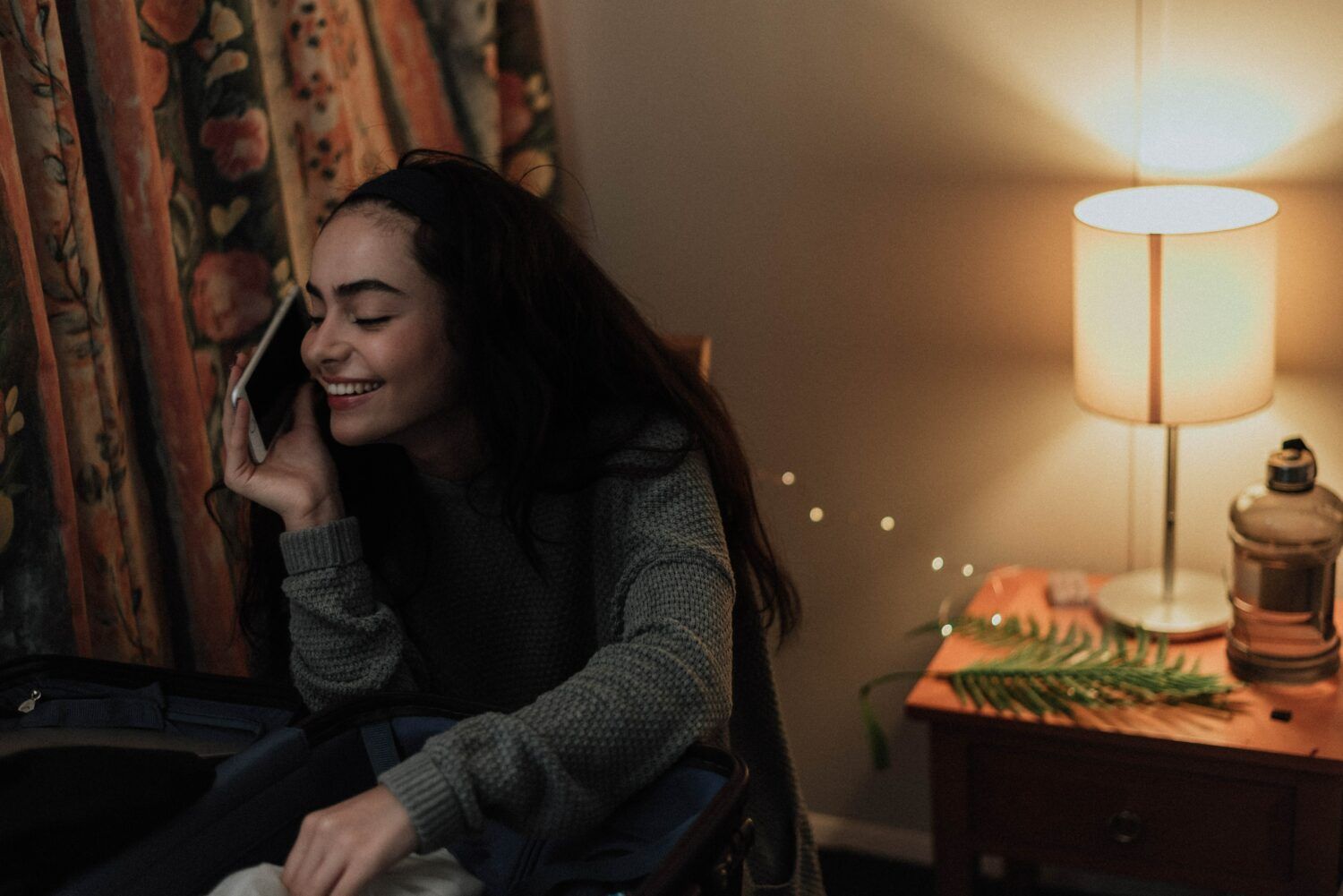 Person smiling on a phone call beside a warm-lit bedside lamp and floral curtains