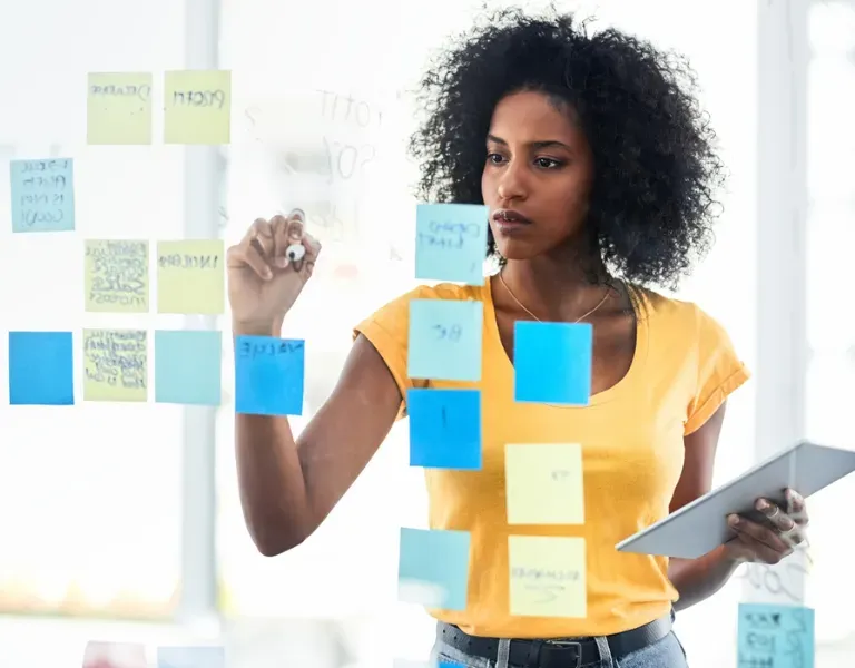Woman writing on glass board with colorful sticky notes while holding a tablet.