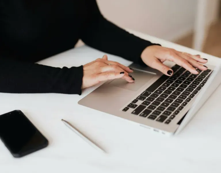 Hands typing on a laptop at a white desk with a smartphone and pen nearby