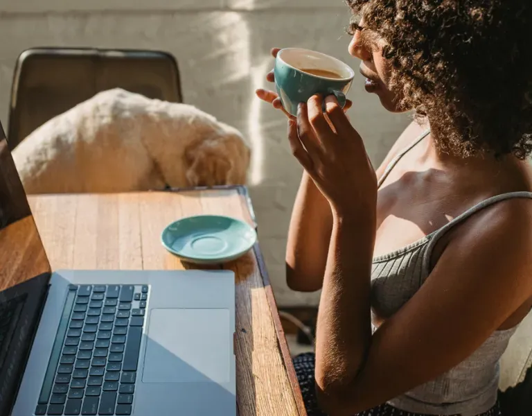 Person sipping from a teal cup beside a laptop at a sunlit table
