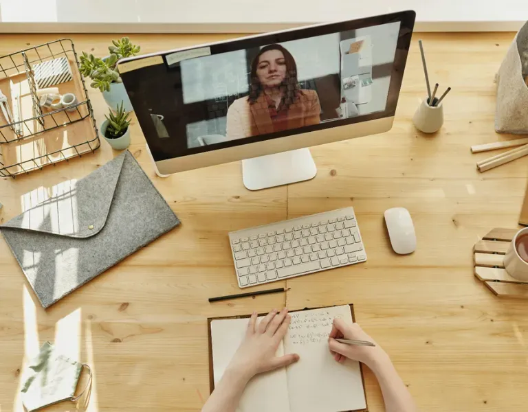 Overhead view of a desk with laptop video call, keyboard, notebook, and hands writing