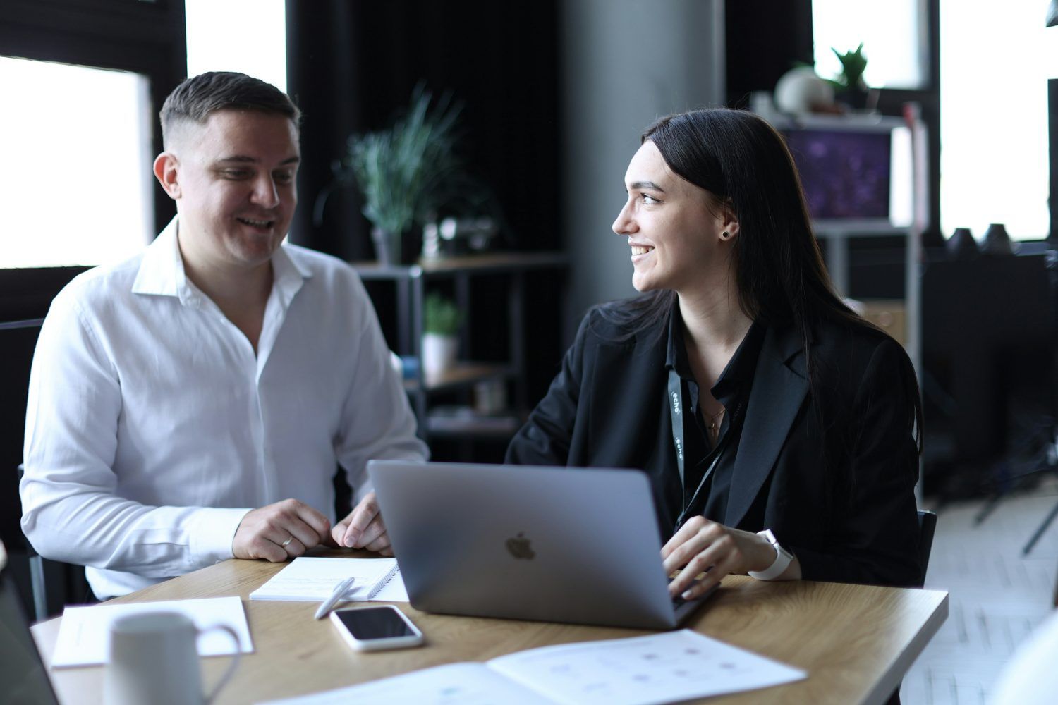 Two coworkers smiling and talking at a table with a laptop in a modern office