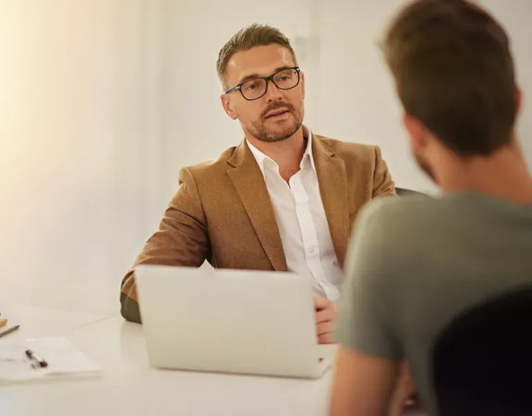 Two people in a meeting across a white desk with a laptop; one man speaks attentively in a bright office