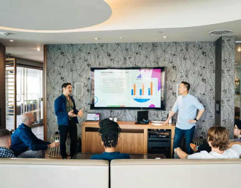 Team meeting in a lounge, with presenters discussing a chart on a large screen.
