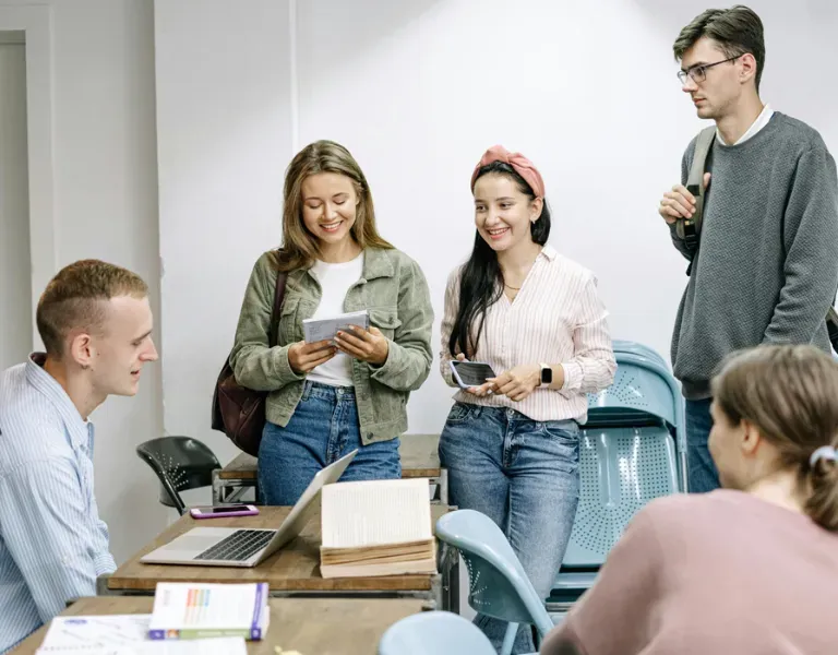 Small group standing and sitting around a table, chatting and using phones in a bright classroom.