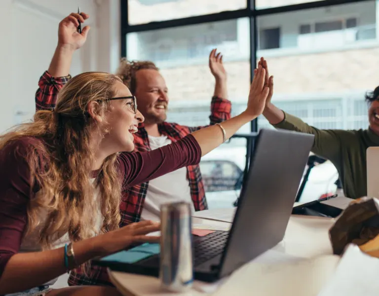 Colleagues in an office high-five during a meeting, smiling beside laptops and a coffee cup