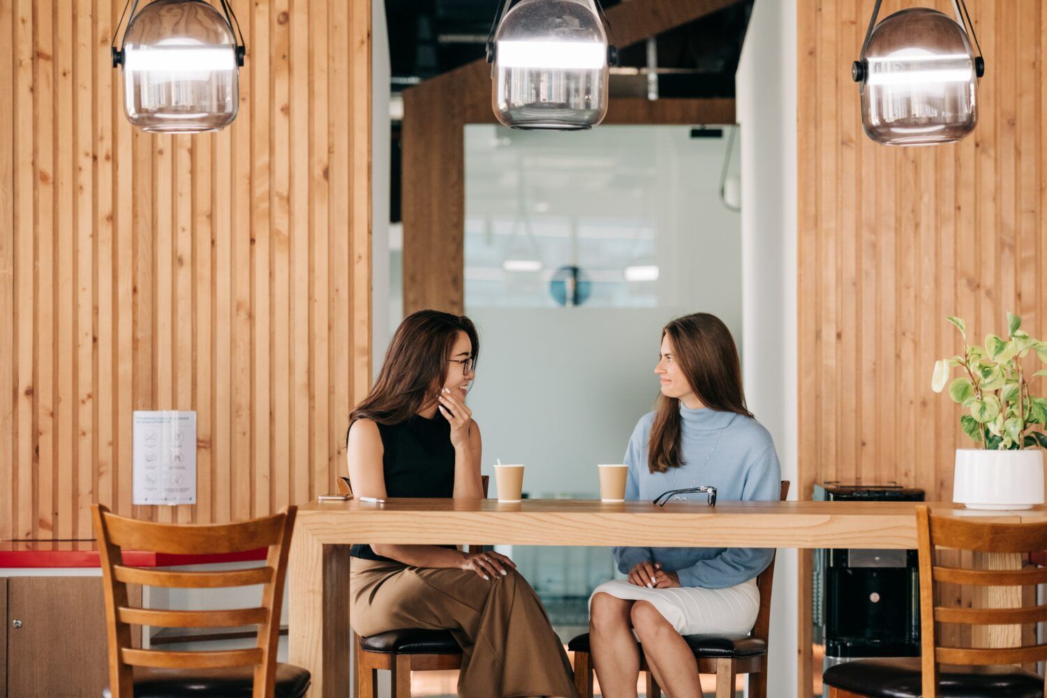 Two people seated at a wooden table in a cozy cafe, facing each other under hanging lights.