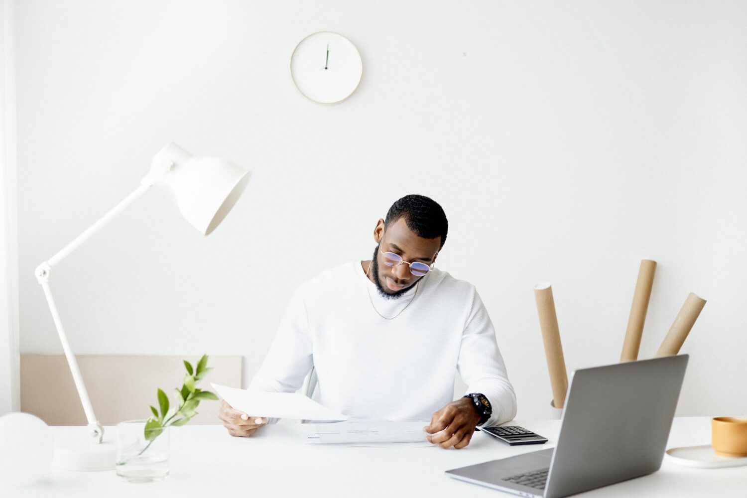 Man in white shirt working at a desk with laptop, papers, lamp, plant, and wall clock